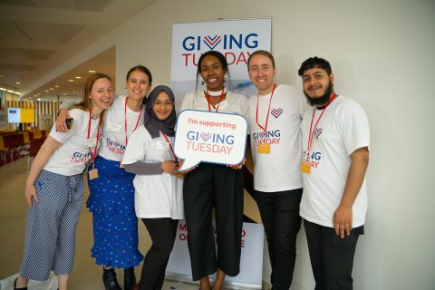 A group of 6 individuals wearing Giving Tuesday t-shirts. The woman in the middle is holding a prop that says "I'm Supporting Giving Tuesday."