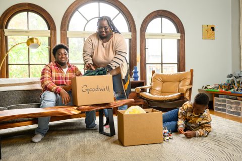 Two parents and a young boy fill up two cardboard boxes labeled "Goodwill" with household items.