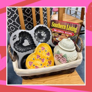 Galentine's gift ideas: a basket full of baking-themed thrifted items including a heart shaped cake pan, heart shaped plates, a floral ceramic cookie jar, and a vintage Southern Living cookbook.