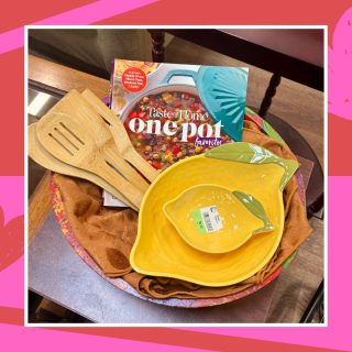 A Galentine's gift basket full of cooking-themed thrifted items including a a fruit bowl, lemon-shaped mixing bowls, wooden cooking utensils, and a one pot cookbook.