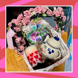 A Galentine's gift basket full of thrifted items catered to a whimsical or cottagecore style including a candle, floral glassware, and a teapot.