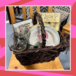 A Galentine's gift basket full of thrifted items including wine glasses, wine glass charms, wine and cocktail-themed plates, and an 'Eat, Pray, Love' DVD.