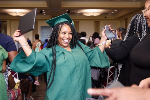 Baltimore Excel Center graduate in green cap and gown celebrates with raised arms at graduation ceremony