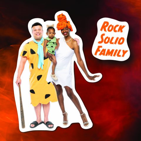 A DIY family Halloween costume based on the Flintstones cartoon TV show. Two parents are wearing dresses and tunics based on Fred and Wilma, while the mother holds a baby wearing a DIY costume to match the Flintstones baby BammBamm.